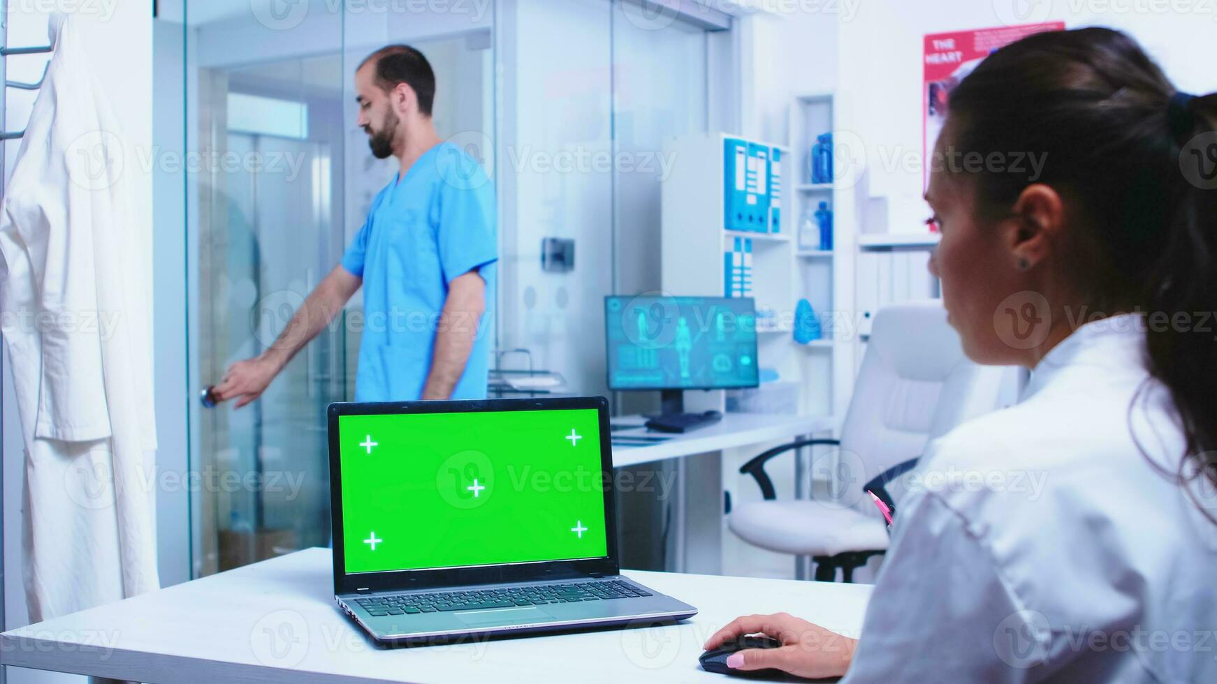 Medical worker in white coat using laptop with green screen in hospital cabinet. Nurse wearing blue uniform opening glass door. Medic wearing uniform using notebook with chroma key on display in medical clinic. photo