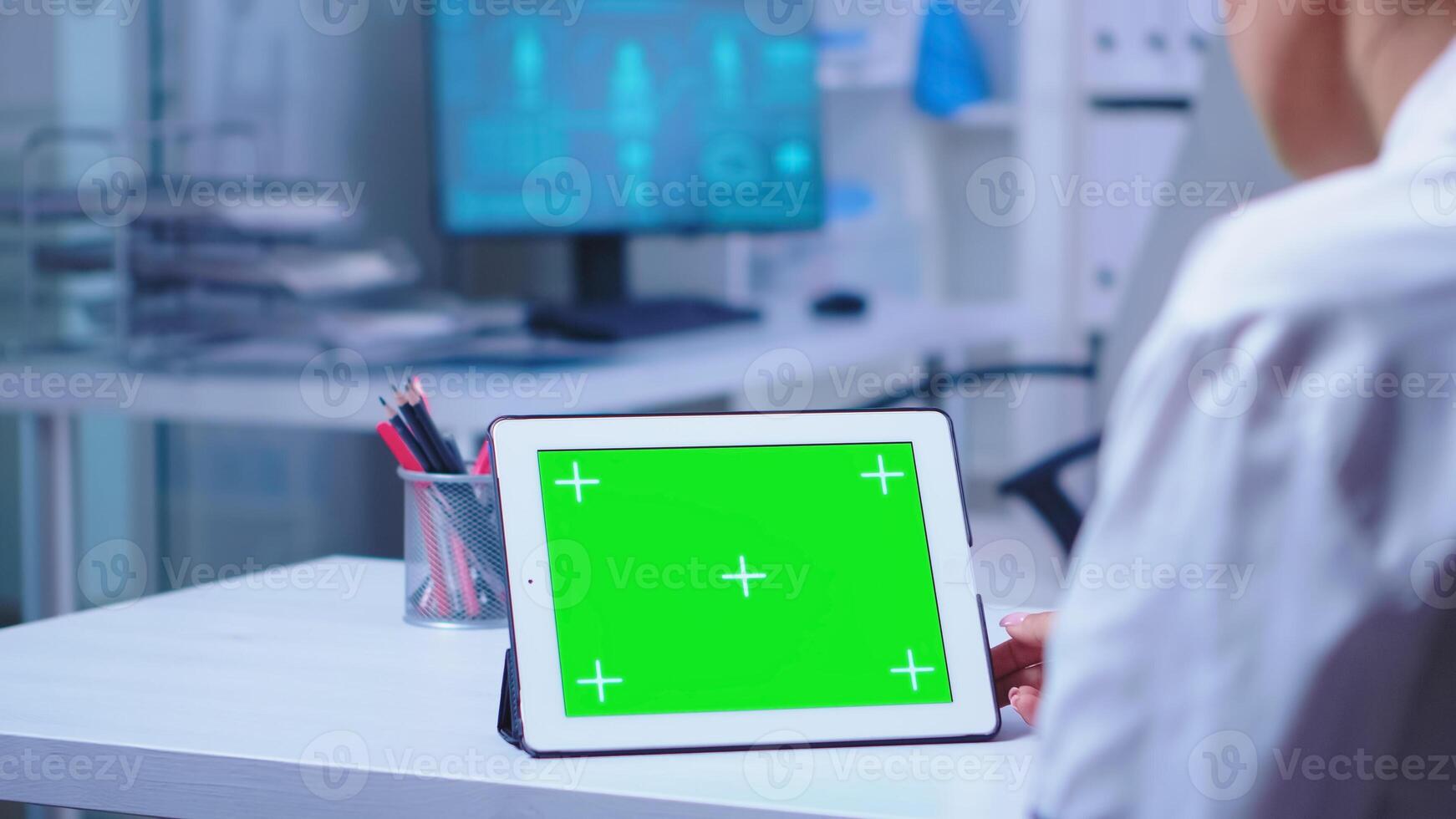 Tablet pc with green screen in hospital cabinet used by doctor wearing white coat. Nurse in blue uniform entering in clinic. Doctor in health clinic working on tablet computer with replaceable screen doing medicine research. photo