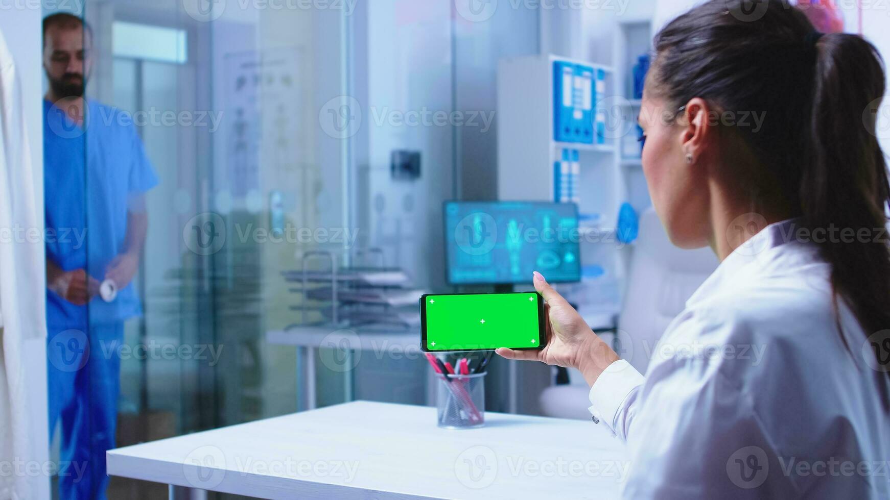 Doctor checking patient results on smartphone with green screen in hospital cabinet. Nurse in blue medical uniform closes glass door. Healthcare specialist in hospital cabinet using smartphone with mockup. photo