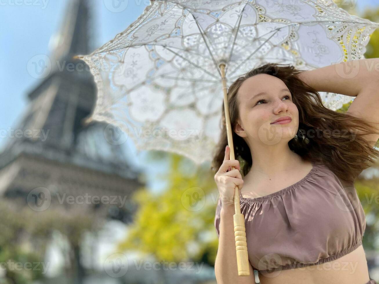 un hermosa joven niña en un verano vestir soportes en contra el fondo de el eiffel torre con un cordón paraguas, ella elevado su mano y como Si tomando apagado allí es un sitio para publicidad viaje agencia foto