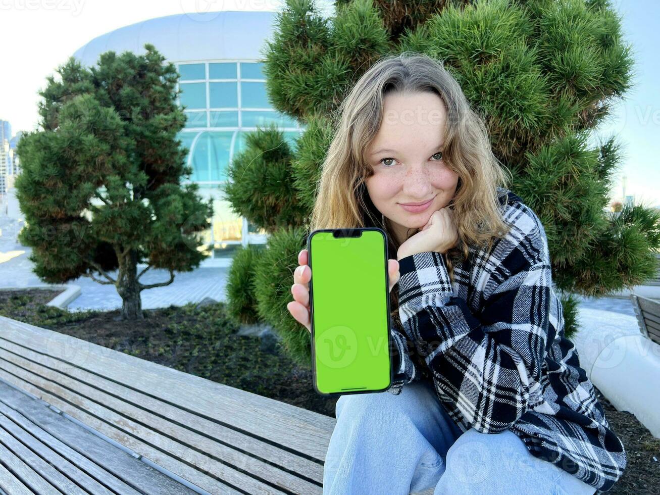 A young woman sits with a phone turned to the camera with a green screen Vancouver Porto Canada Place Coniferous trees are in the background. She is wearing a black and white checkered blouse. photo