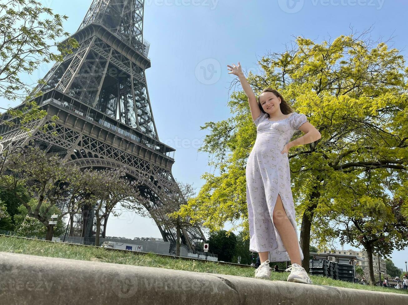 un alegre adolescente niña soportes en contra el fondo de el eiffel torre en un hermosa vestido, ella elevado su mano, usted lata anunciar un viaje agencia viaje a París Francia foto