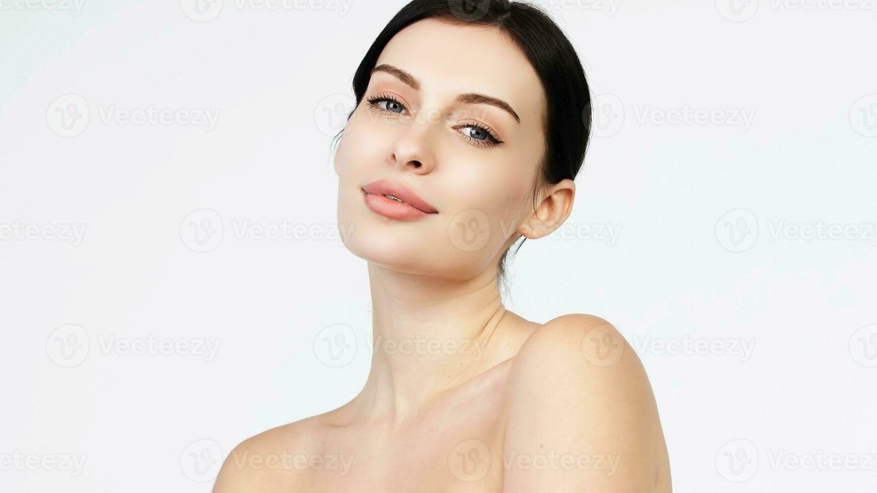 Close-up beauty portrait of young woman with smooth healthy skin, she gently touches her face with her fingers on light grey background and smiles. Skincare concept photo