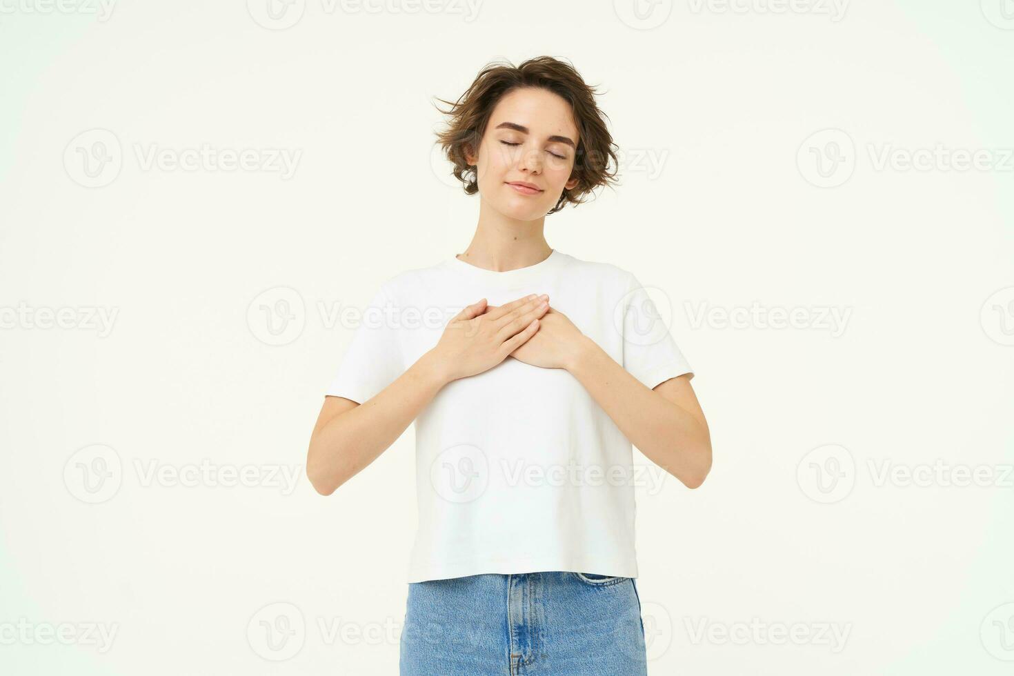 Portrait of woman expressing care, self-love and caring emotions, holds hands on chest, looking with smile at camera, isolated over white background photo