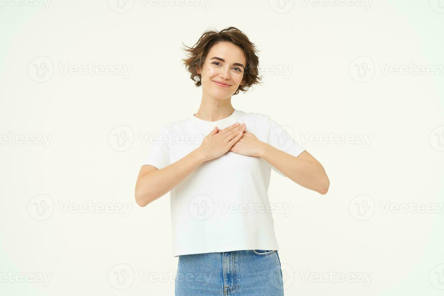 Portrait of brunette woman with hands on chest, looks with care and love, express gratitude, self-care and heart-warming feelings, stands over white background photo