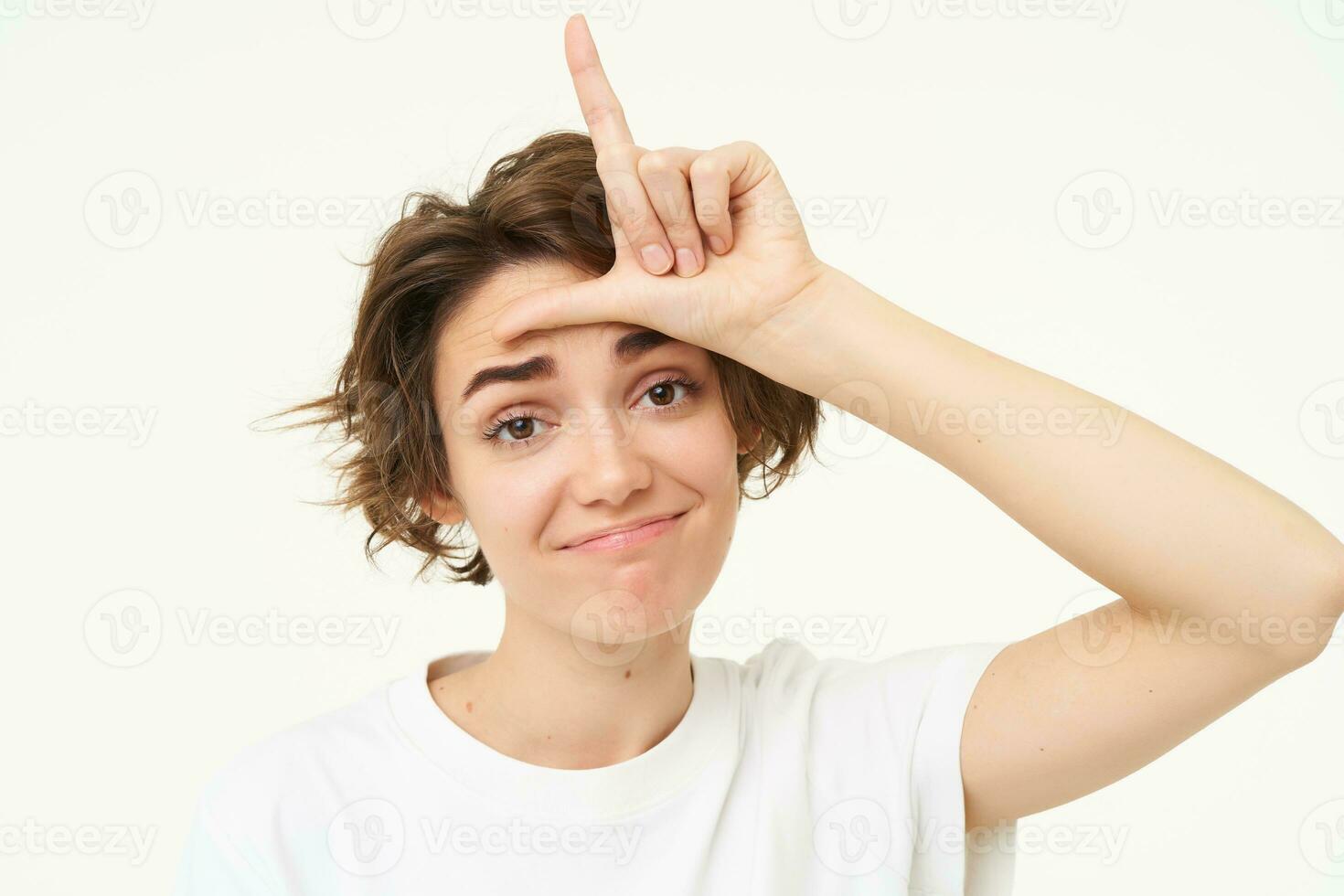 Close up of woman shows loser gesture, l letter on forehead and smiling, mocking, makes fun of someone, standing over white background photo
