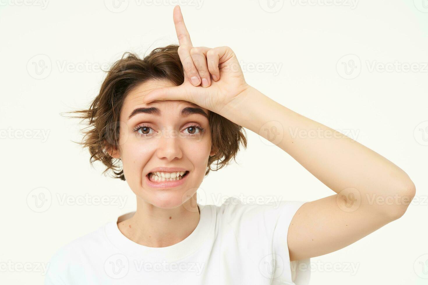 Close up of young woman making fun of friend, smiling and showing loser gesture, l letter on forehead, standing over white background photo