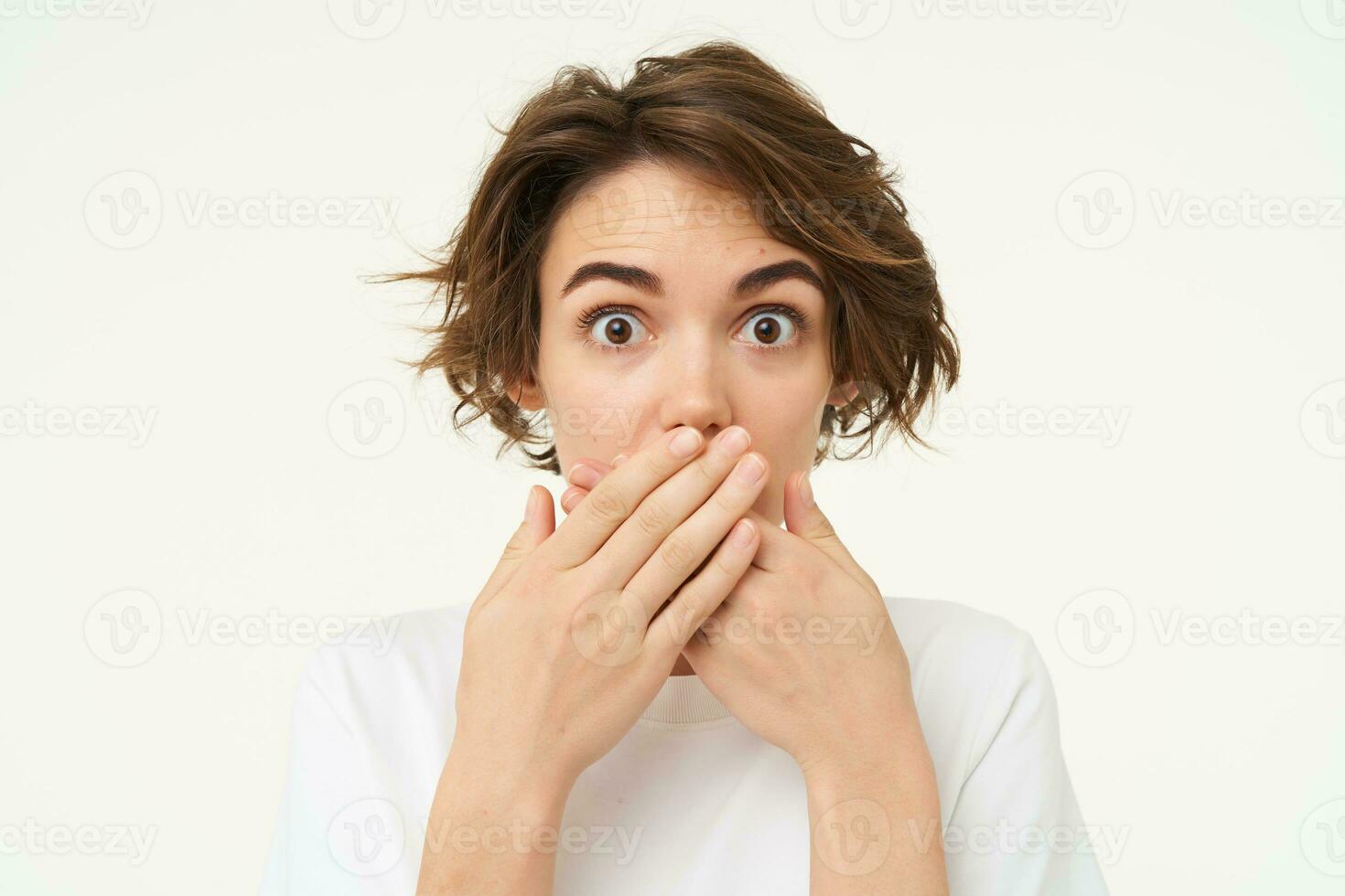 Image of brunette woman shuts her mouth, looks surprised, puts hands over her lips, stands over white background photo