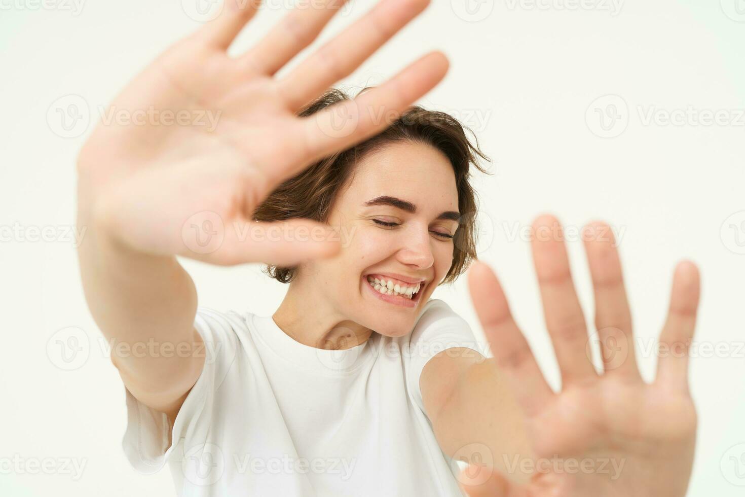 Close up of happy brunette woman extending her hands, stretching, covering herself from camera, laughing and smiling, standing over white background photo