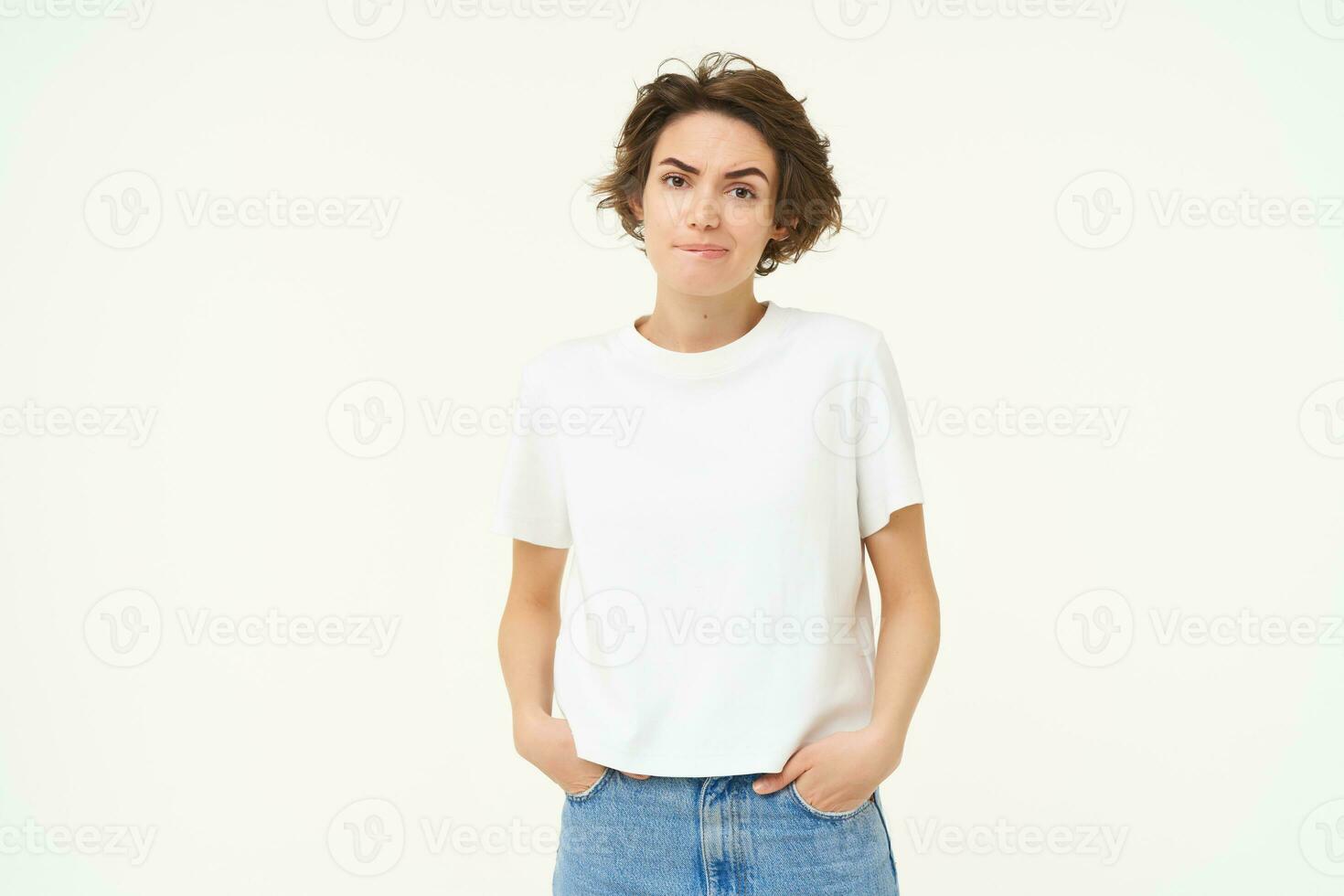 Portrait of woman looking reluctant and unconfident, frowning, holding hands in pockets, standing doubtful, feeling unsure, white studio background photo