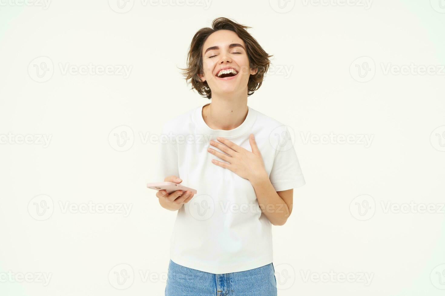 Portrait of young brunette woman texting, sending a message on telephone, using mobile phone and smiling, standing over white studio background photo