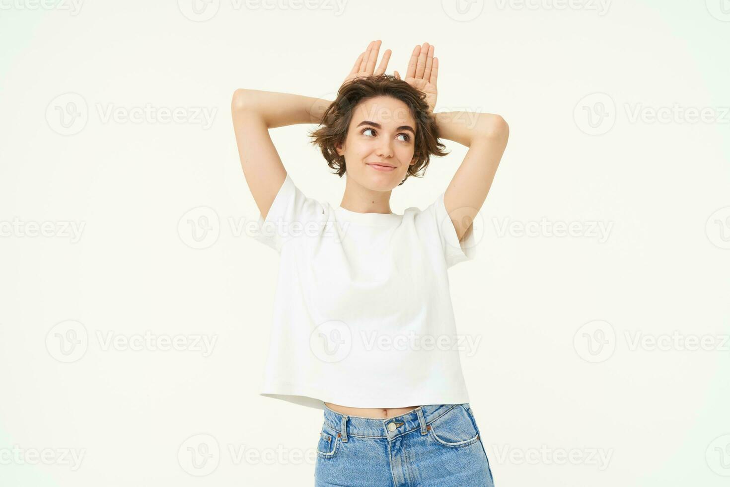 Portrait of brunette girl, makes silly face, shows animal ears on head, posing over white studio background photo