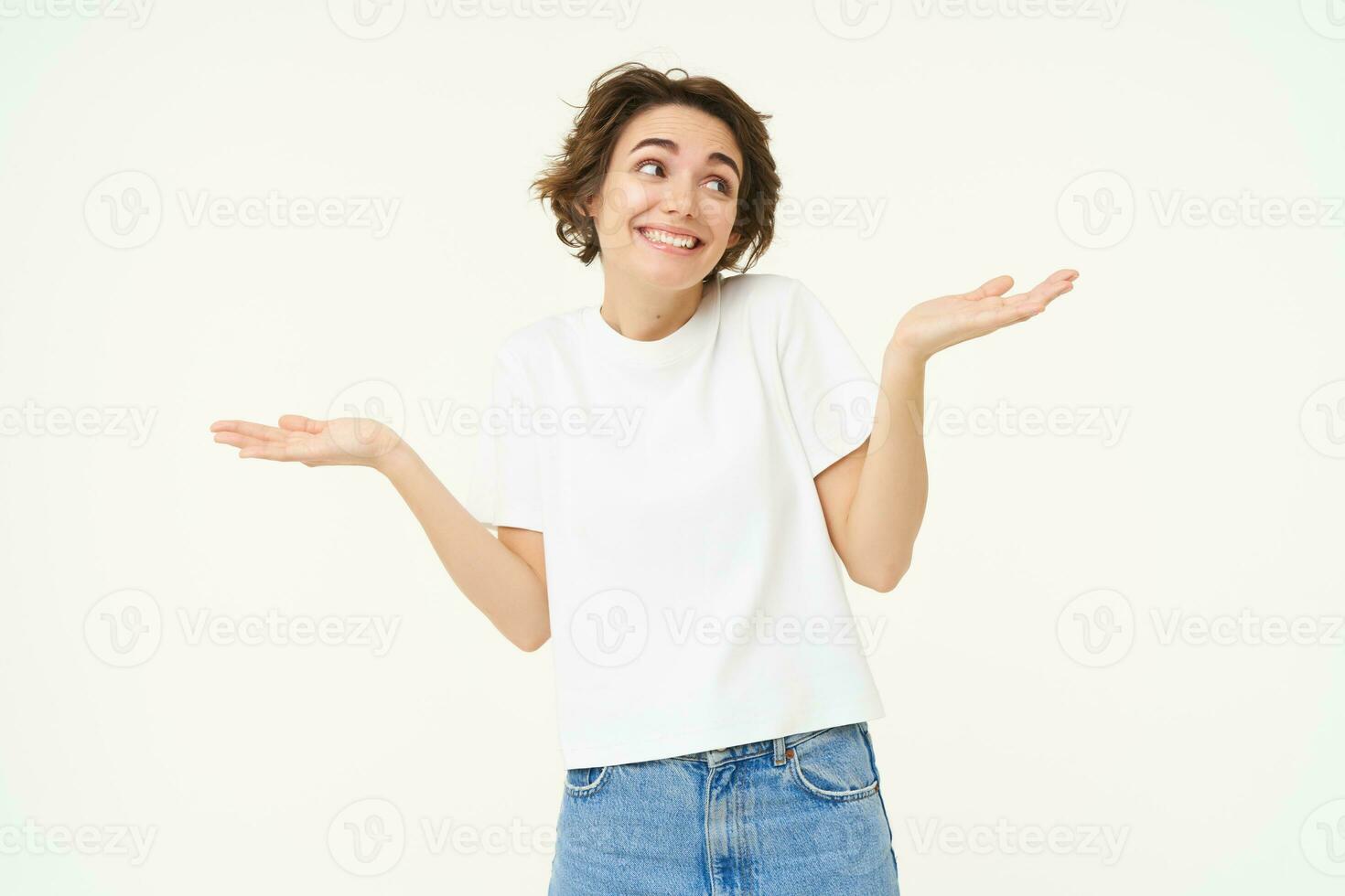 Portrait of girl with short hair, shrugs shoulders, looks unaware, clueless, doesnt know what to do, stands over white studio background photo