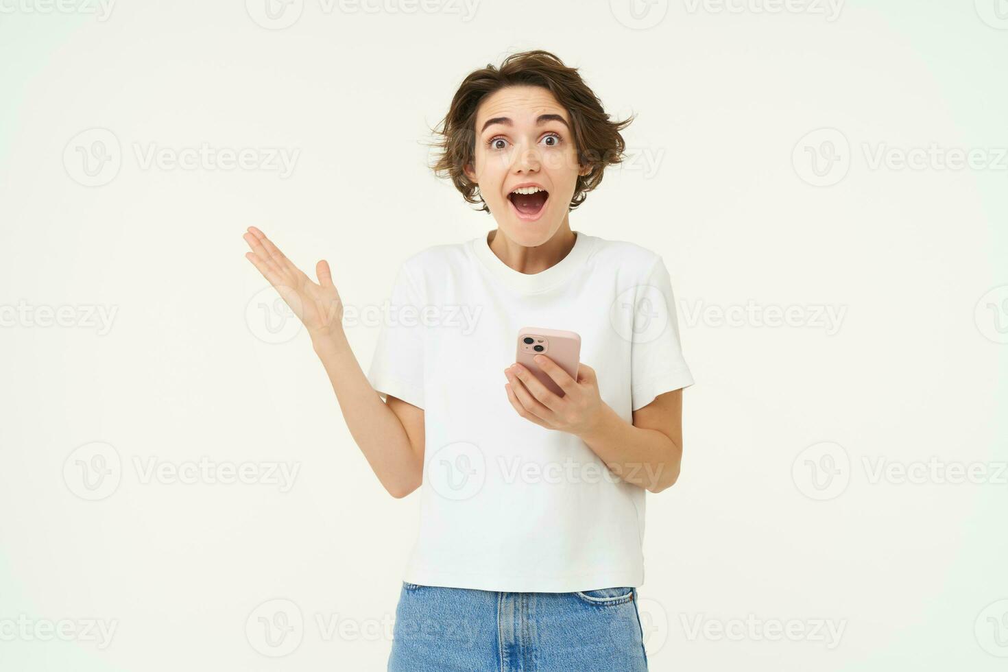 Portrait of brunette girl expresses amazement and surprise, holds smartphone, reacts to amazing advertisement on mobile phone, standing over white background photo