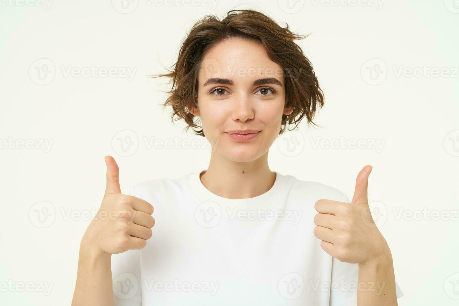 Close up of happy brunette woman, shows thumbs up, approves something, recommends, gives positive feedback, stands over white background photo