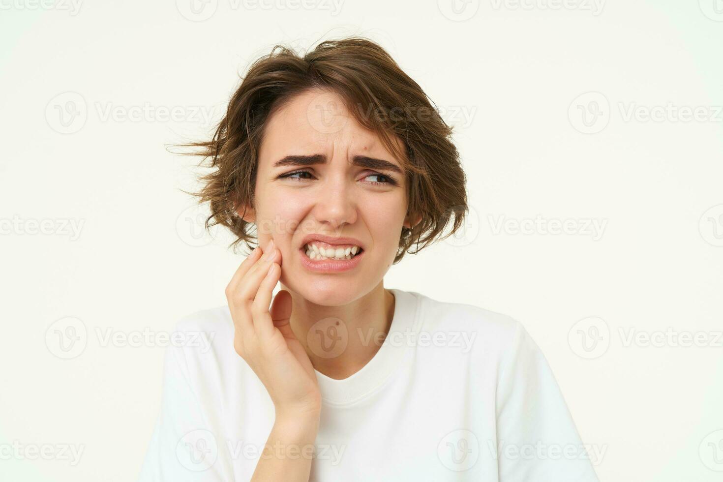 Close up of woman has a toothache, touches her teeth and frowns from painful discomfort, stands over white background photo