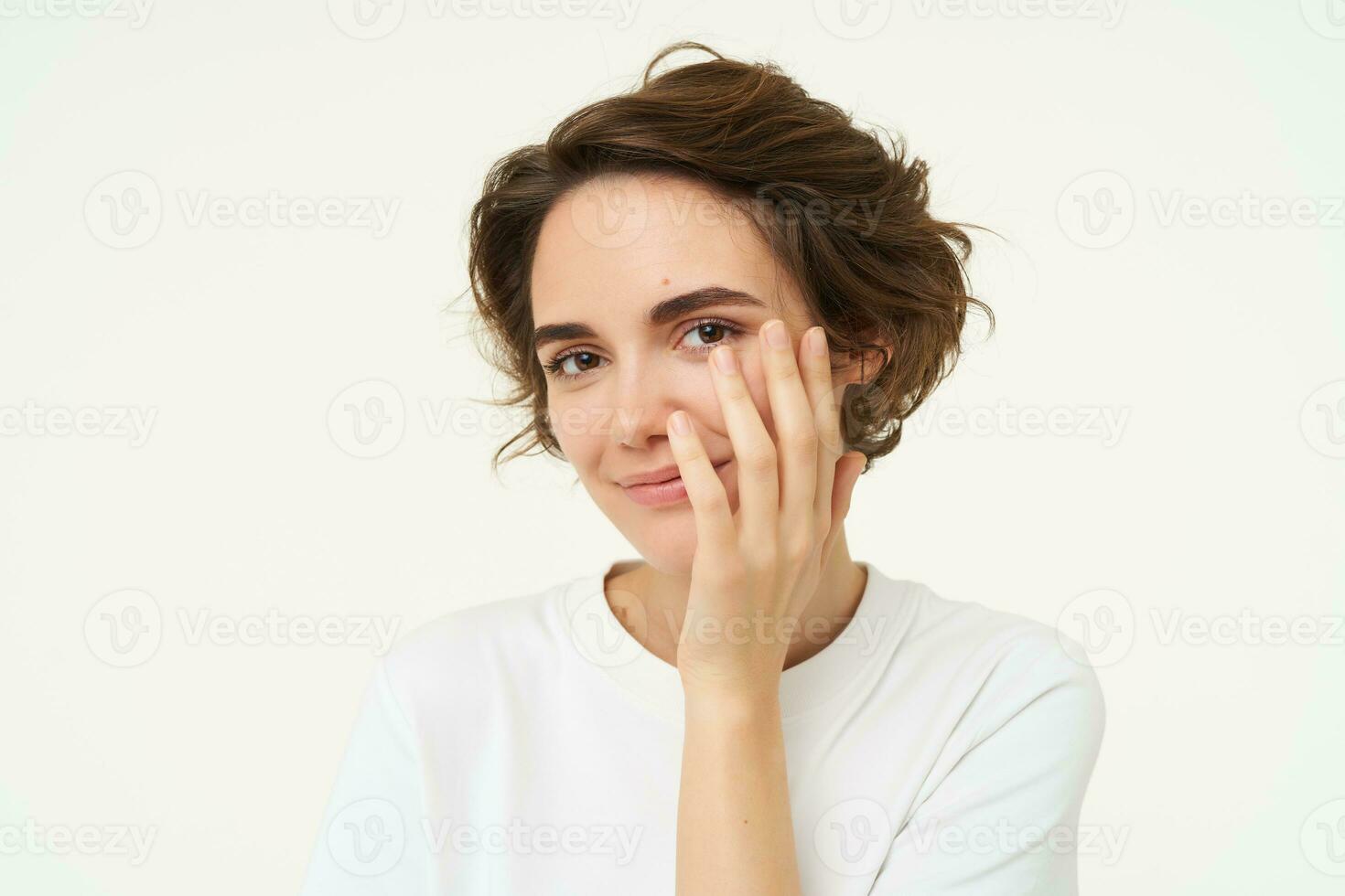 Close up of brunette woman looks shy, covers face with hand and smiles, gazing flirty at camera, stands over white background photo