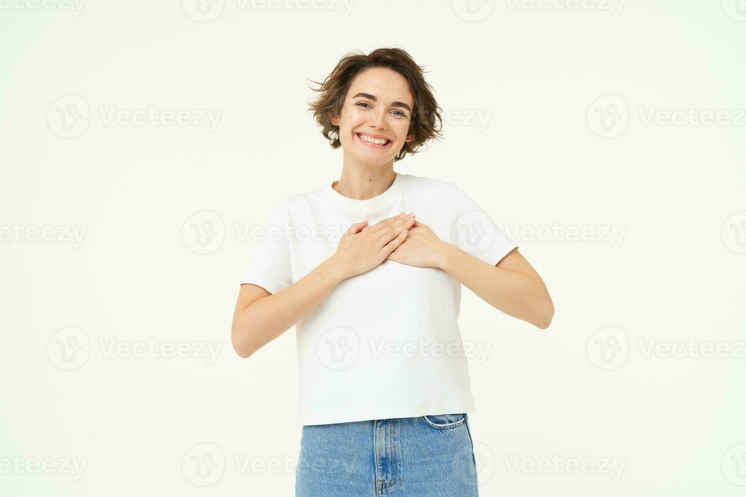 Portrait of woman expressing care, self-love and caring emotions, holds hands on chest, looking with smile at camera, isolated over white background photo