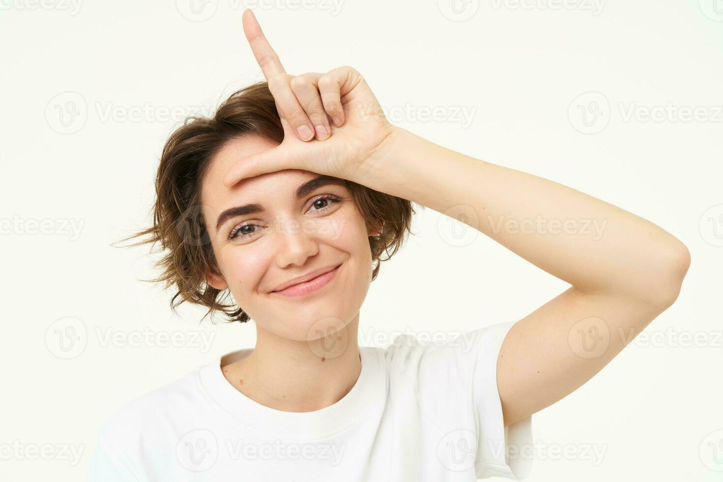 Close up of young woman making fun of friend, smiling and showing loser gesture, l letter on forehead, standing over white background photo