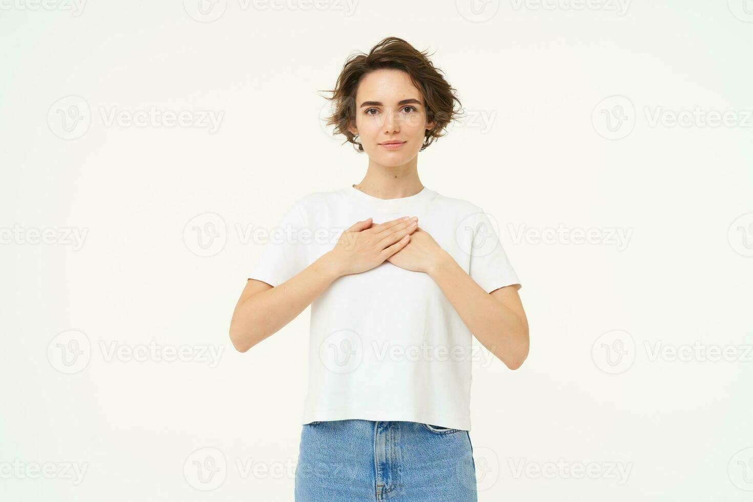 Portrait of brunette woman with hands on chest, looks with care and love, express gratitude, self-care and heart-warming feelings, stands over white background photo
