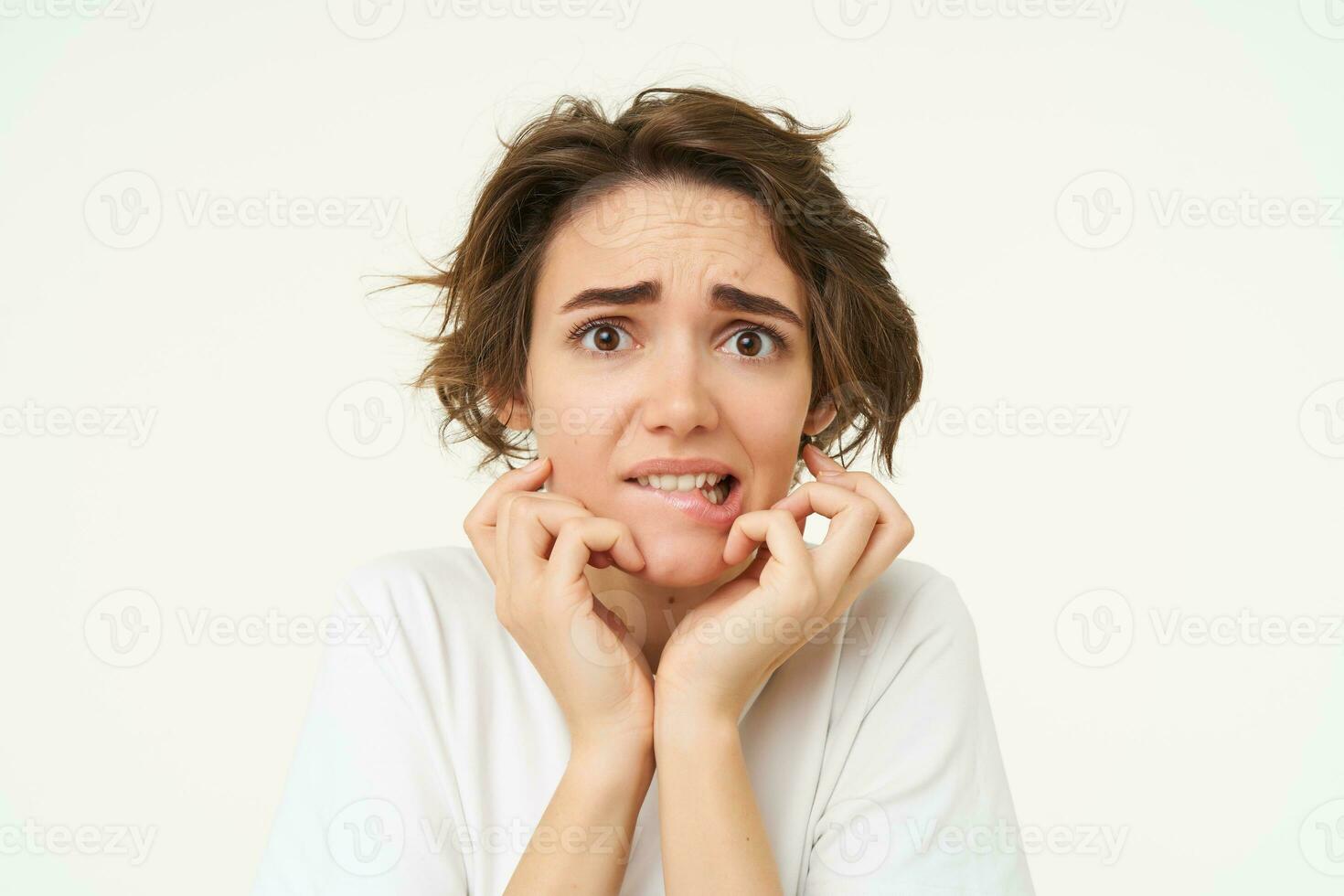 Close up of scared brunette woman, shaking from fear, looking concerned and frightened, standing over white studio background photo