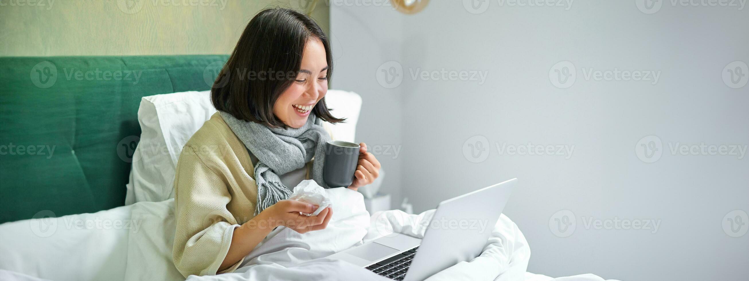 Portrait of asian sick girl, lying in bed with cold, having flu, talking to laptop, video chat, working from home on remote while on sick leave photo