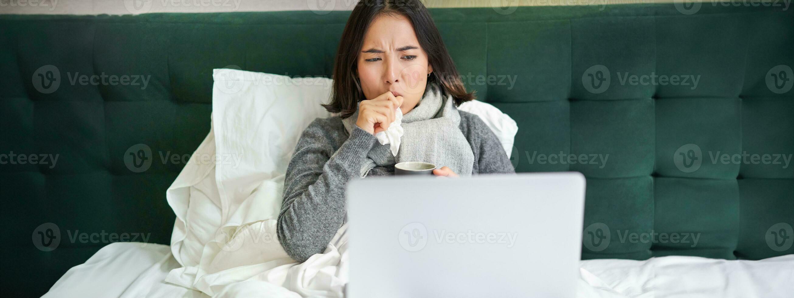 Portrait of sick asian woman watching videos on laptop, staying in bed and coughing, catching cold photo