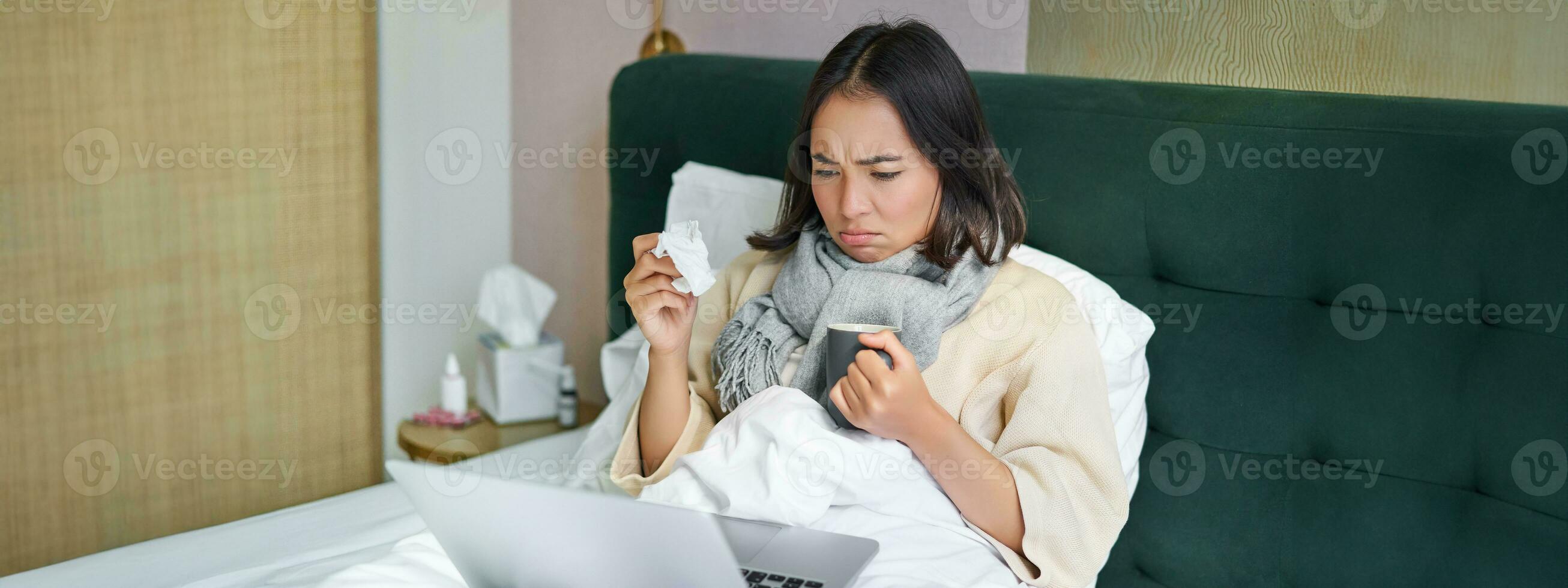 Portrait of asian girl sneezing and wiping runny nose, feeling sick, staying in bed with influenza, looking at laptop photo