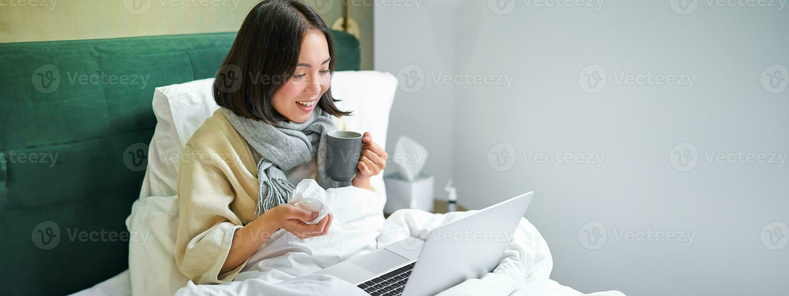 Portrait of asian sick girl, lying in bed with cold, having flu, talking to laptop, video chat, working from home on remote while on sick leave photo