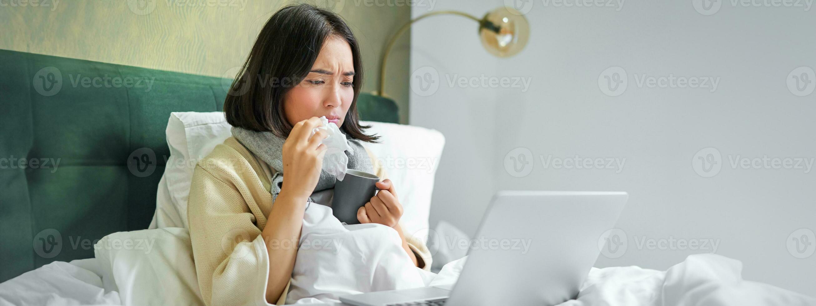 Portrait of asian girl sneezing and wiping runny nose, feeling sick, staying in bed with influenza, looking at laptop photo