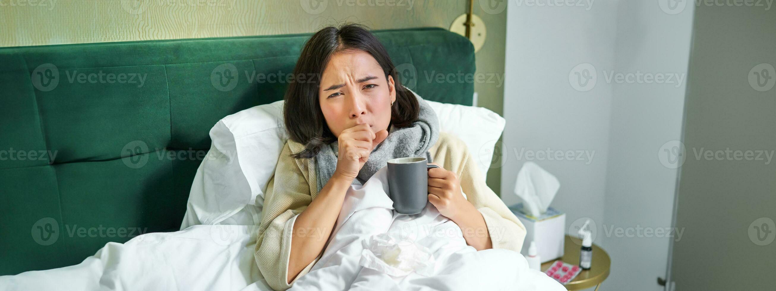 Portrait of couching asian girl in bed, feeling sick, catching cold and staying at home, looking unhealthy, drinking hot tea photo