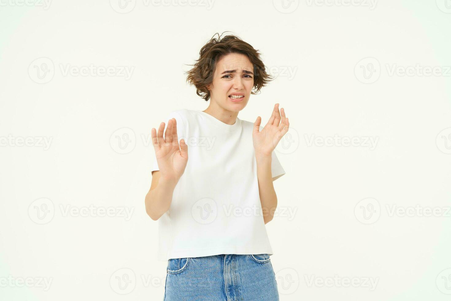 Young reluctant woman steps back, raises hands to refuse, rejects something disgusting, reaction of aversion and dislike, isolated over white background photo