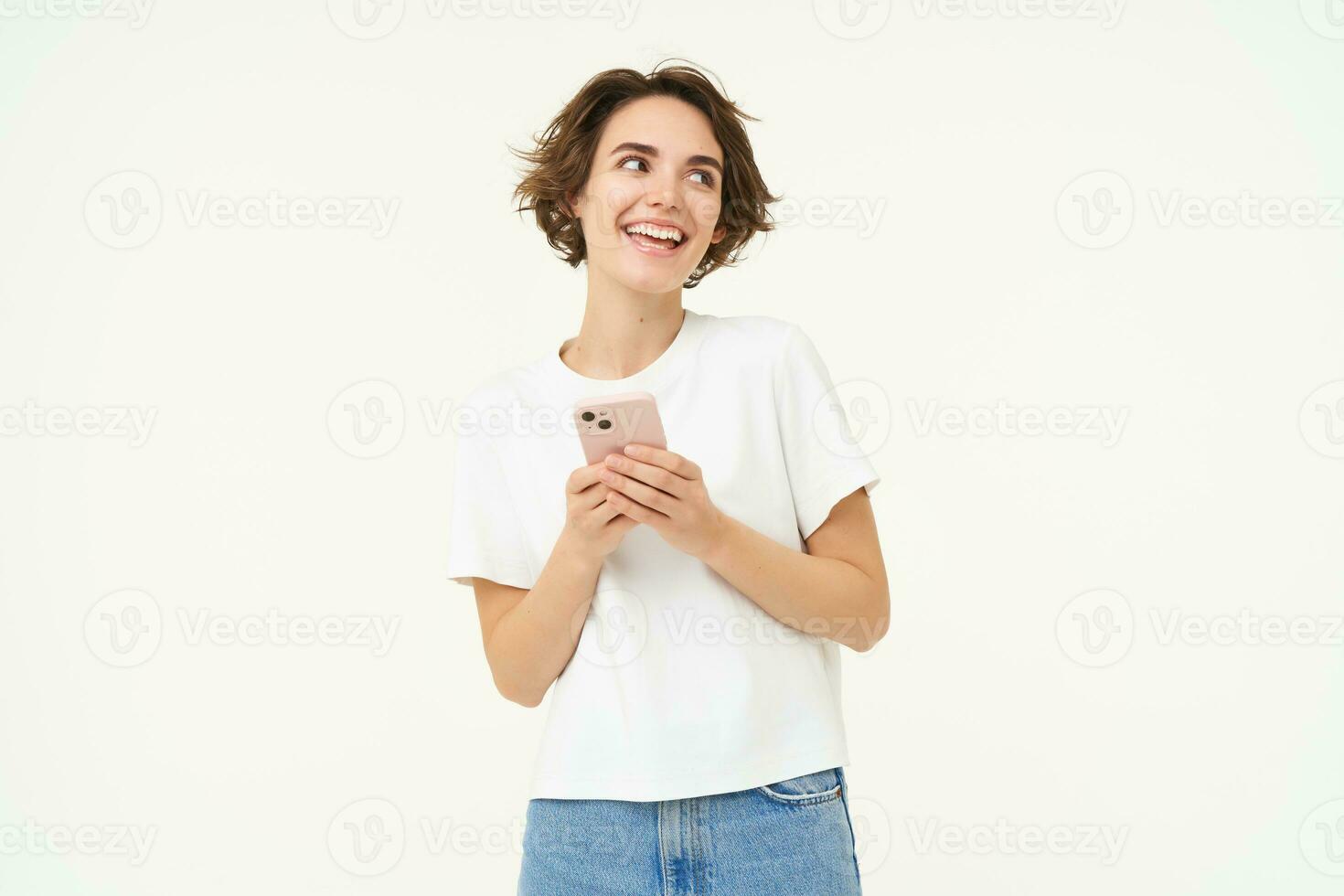 Portrait of young brunette woman texting, sending a message on telephone, using mobile phone and smiling, standing over white studio background photo
