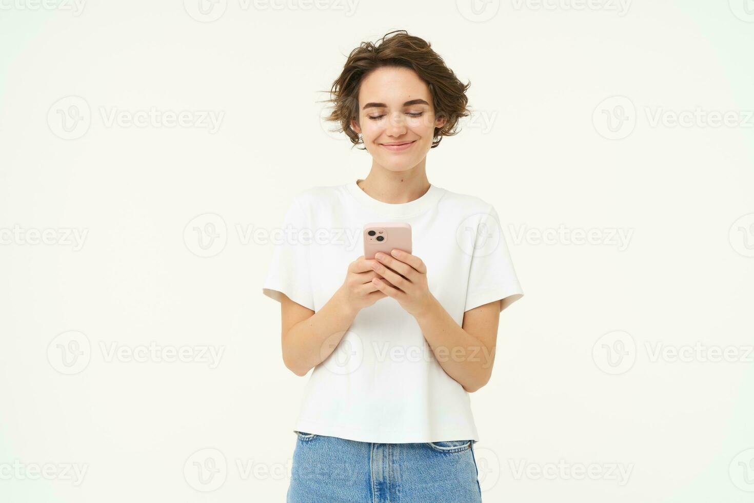 Portrait of young brunette woman texting, sending a message on telephone, using mobile phone and smiling, standing over white studio background photo