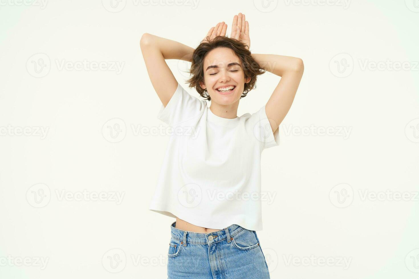 Portrait of brunette girl, makes silly face, shows animal ears on head, posing over white studio background photo