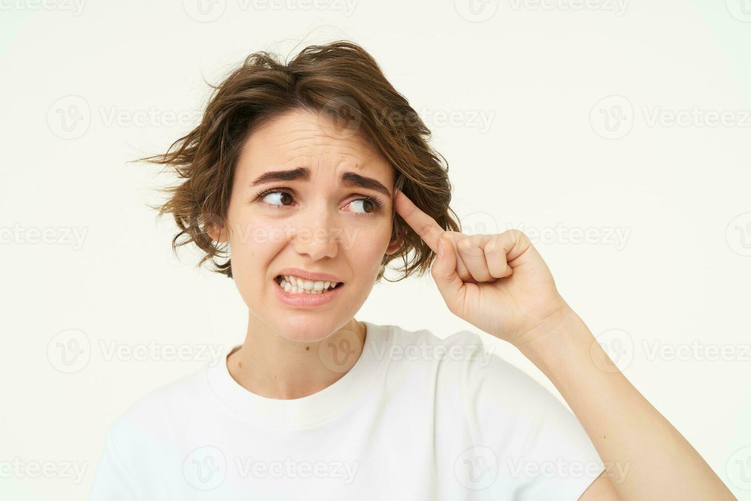 Image of woman cringe, points finger at head, avoids eye contact, thinks someone is crazy, isolated over white background photo
