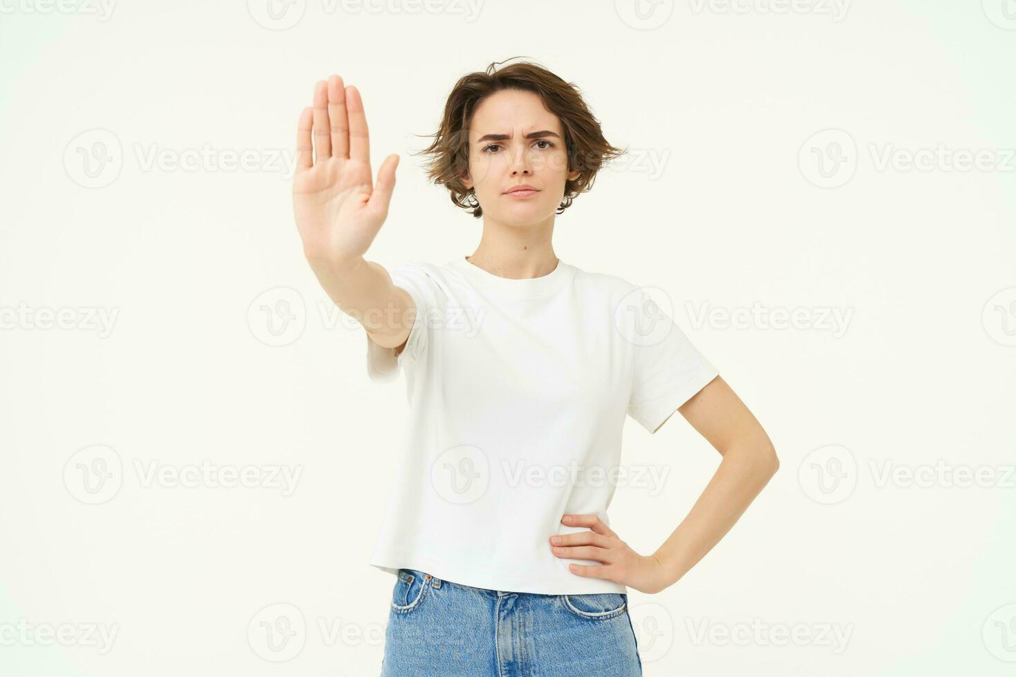 Portrait of serious, confident young woman extends one hand, shows stop, disapproval gesture, looking determined to block way, isolated over white background photo