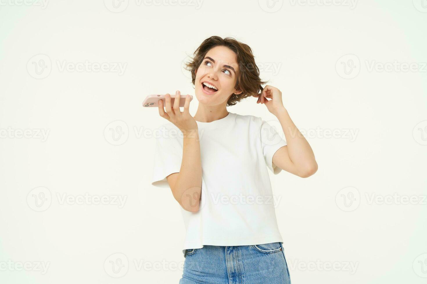 Portrait of chatty woman talking in speakerphone, records voice message on mobile phone and smiling, stands over white background photo