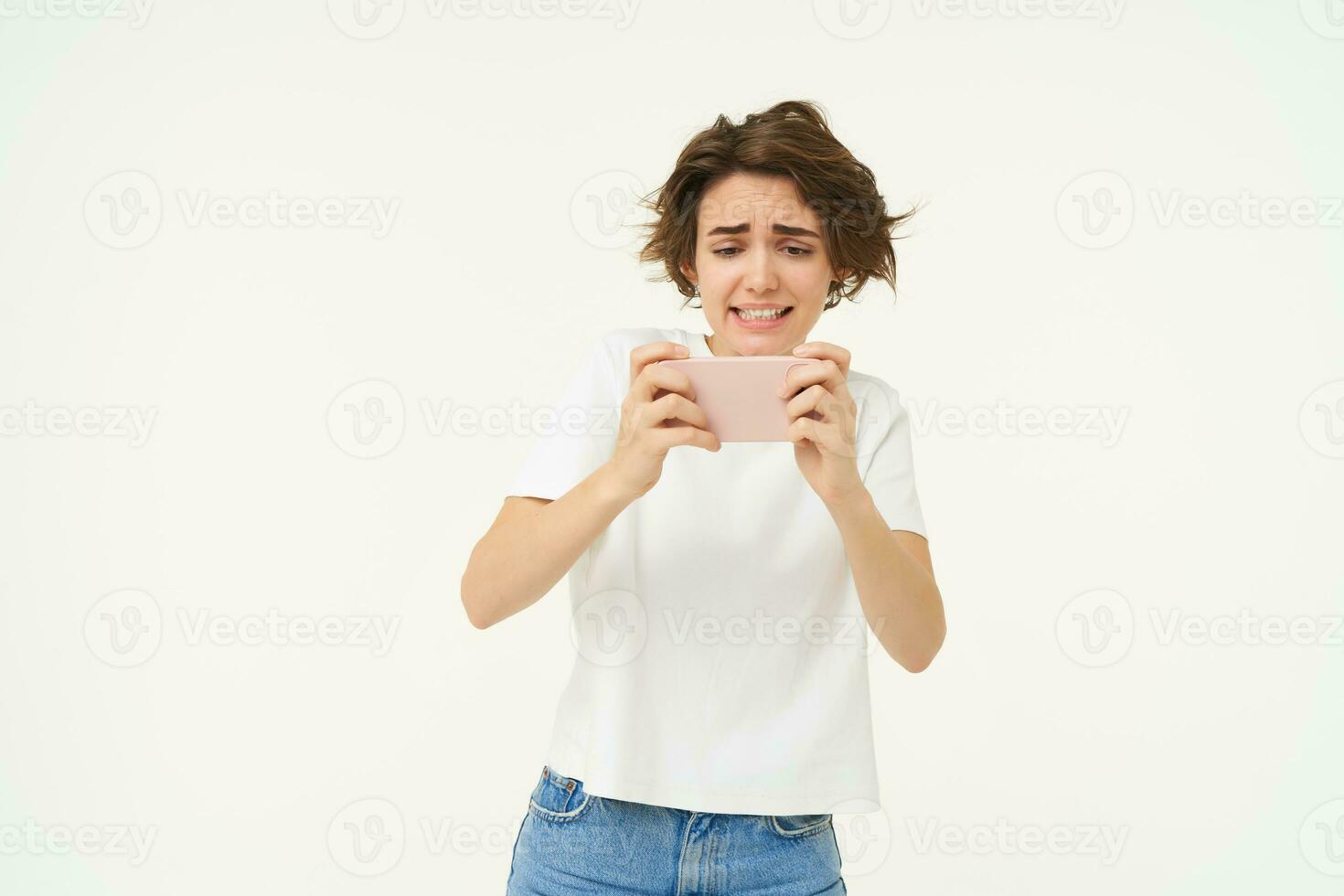 Portrait of woman playing video games on mobile phone, holding smartphone with both hands, standing over white studio background photo