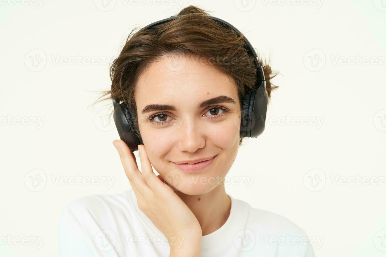 Image of young modern woman in wireless headphones, using earphones to listen to music, standing over white background photo