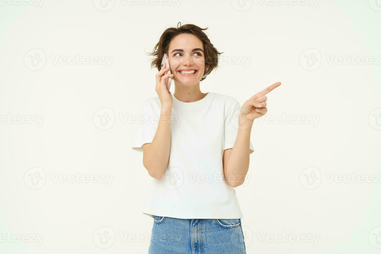 Portrait of friendly, smiling cute girl, pointing finger right, talking on mobile phone, showing smth aside on white copy space, isolated on studio background photo