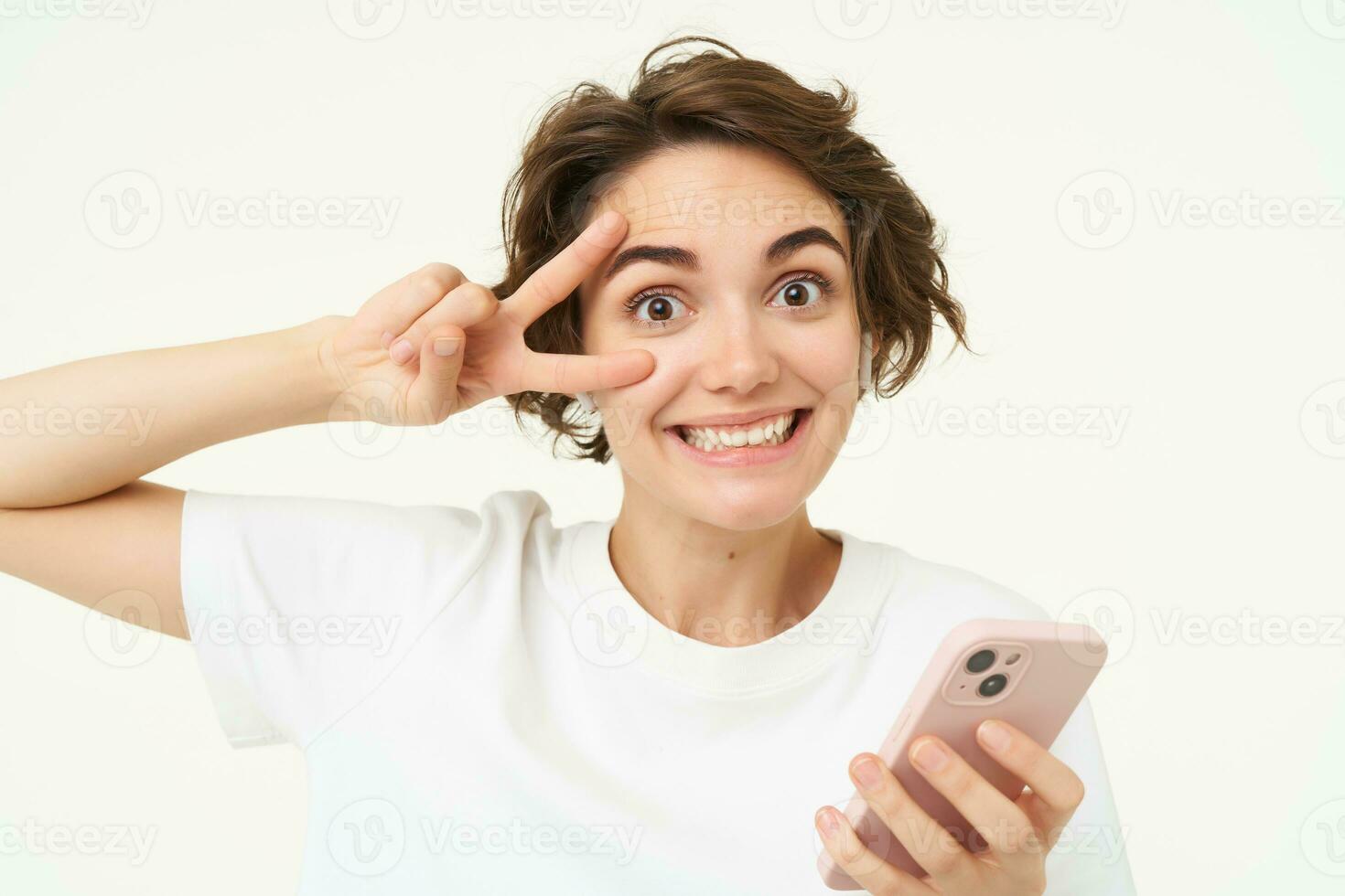 Portrait of young woman shows peace sign, holds smartphone, listens to music in wireless headphones, stands over white background photo