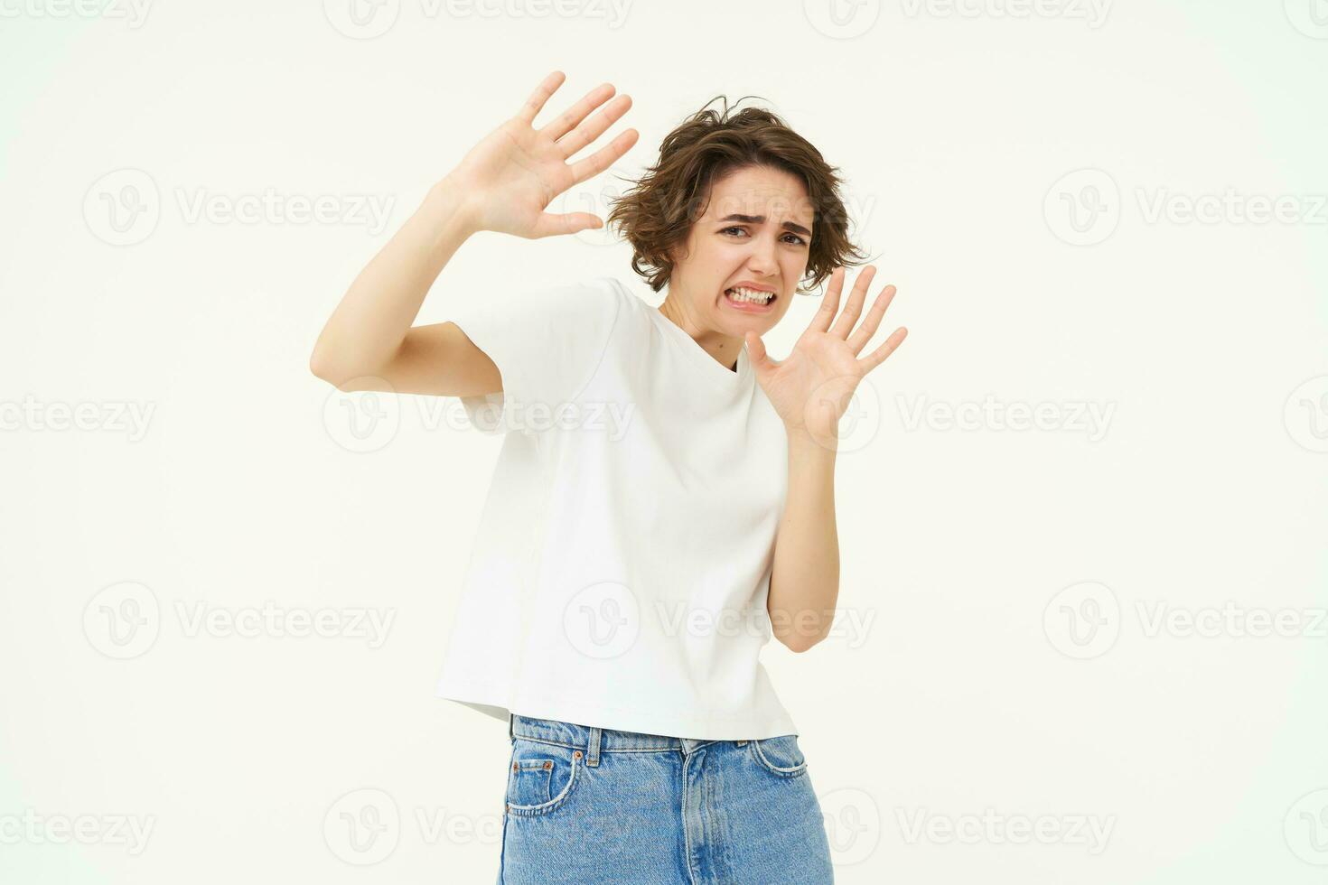 Portrait of woman raising hands in defense and blocking face from something, standing over white background photo