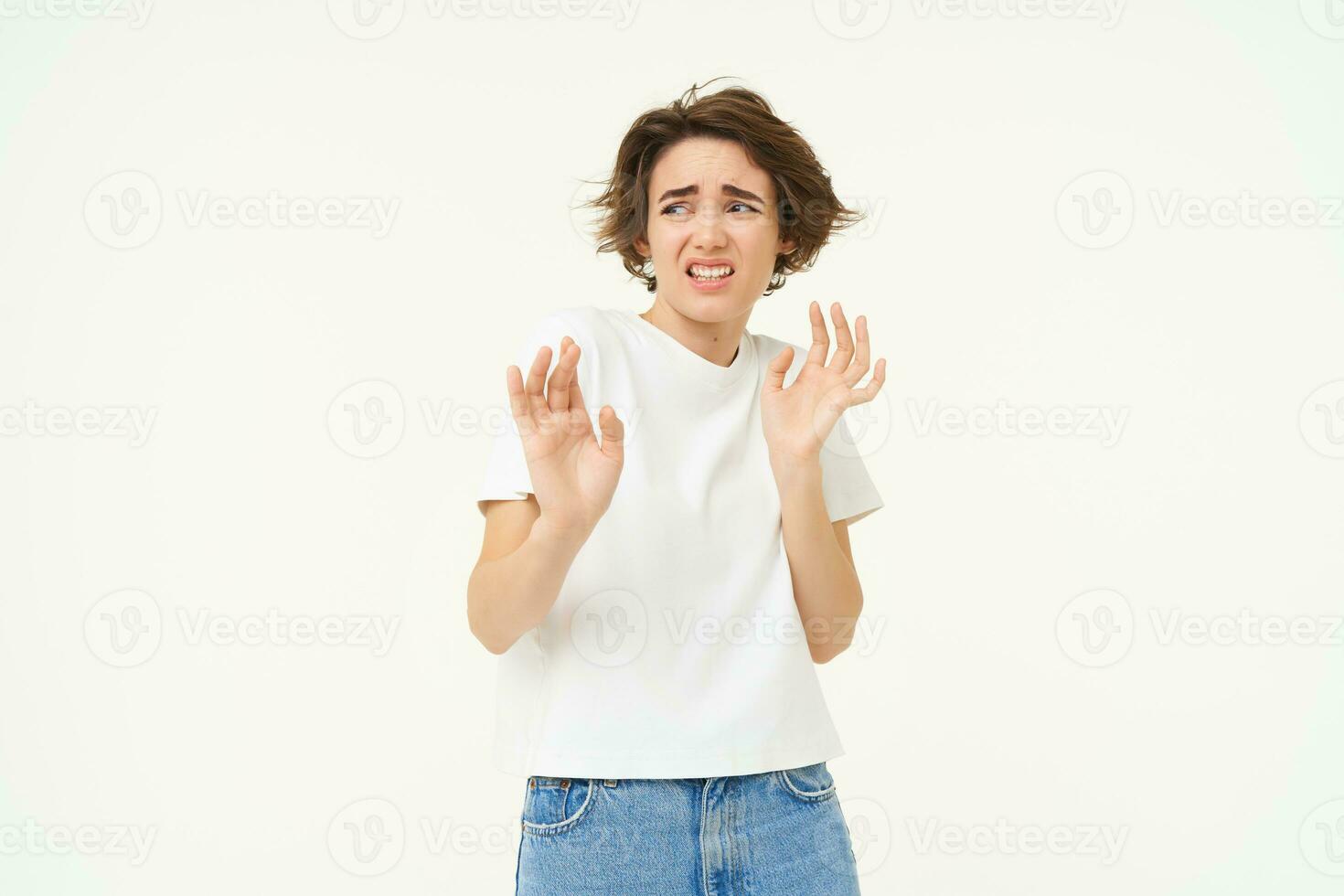 Young reluctant woman steps back, raises hands to refuse, rejects something disgusting, reaction of aversion and dislike, isolated over white background photo
