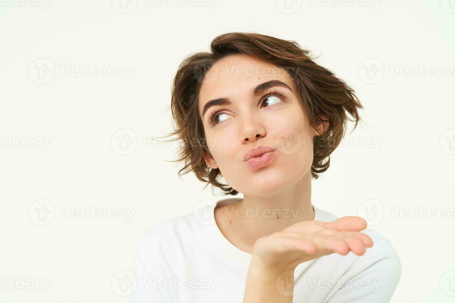 Close up of brunette woman blowing air kiss, mwah, kissing gesture, pucker lips and gazing away, standing over white background photo