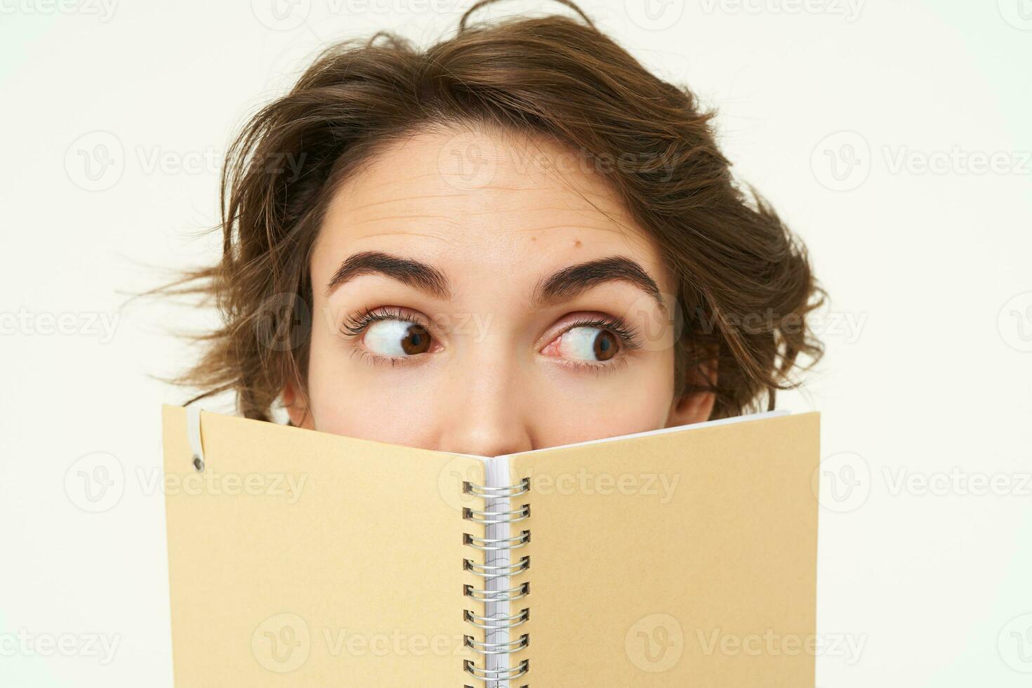 Close up portrait, young woman hiding her face behind planner, holding notebook and looking sideways, side eye, standing over white background photo