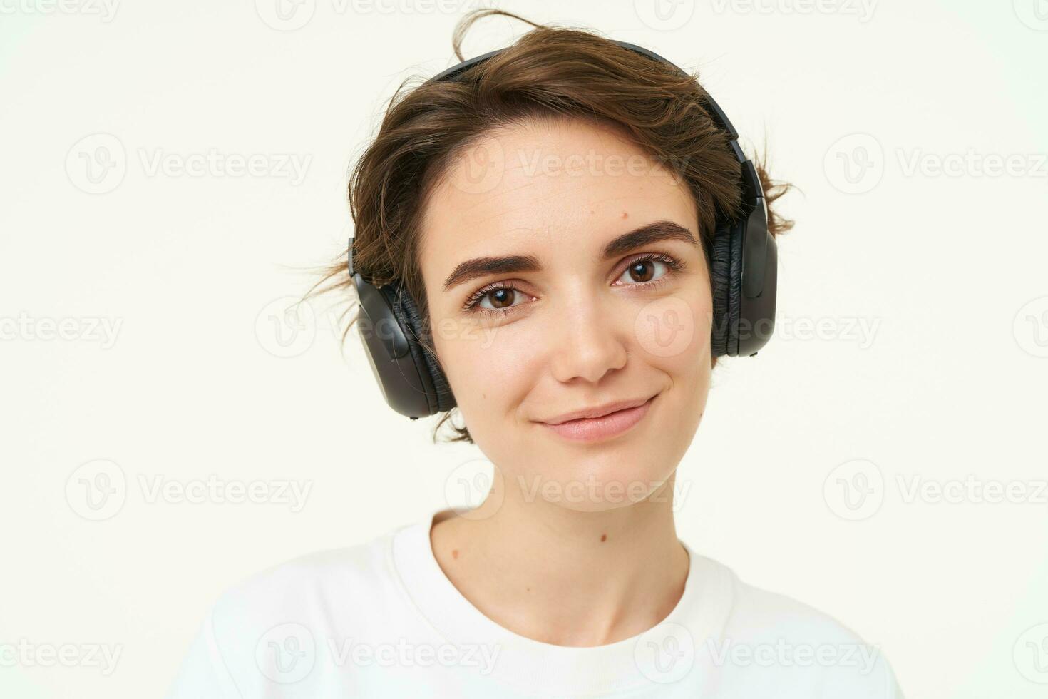 Portrait of young candid woman in wireless headphones, smiling, listening music in earphones, standing over white background photo