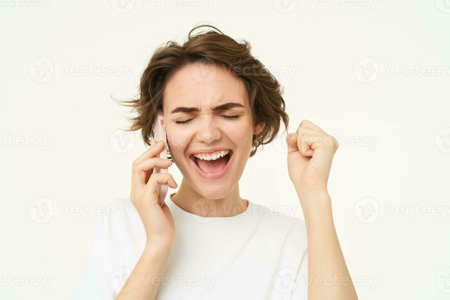 Excited brunette woman celebrating, answers phone call and looking happy, winning, triumphing from great big news, standing over white background photo
