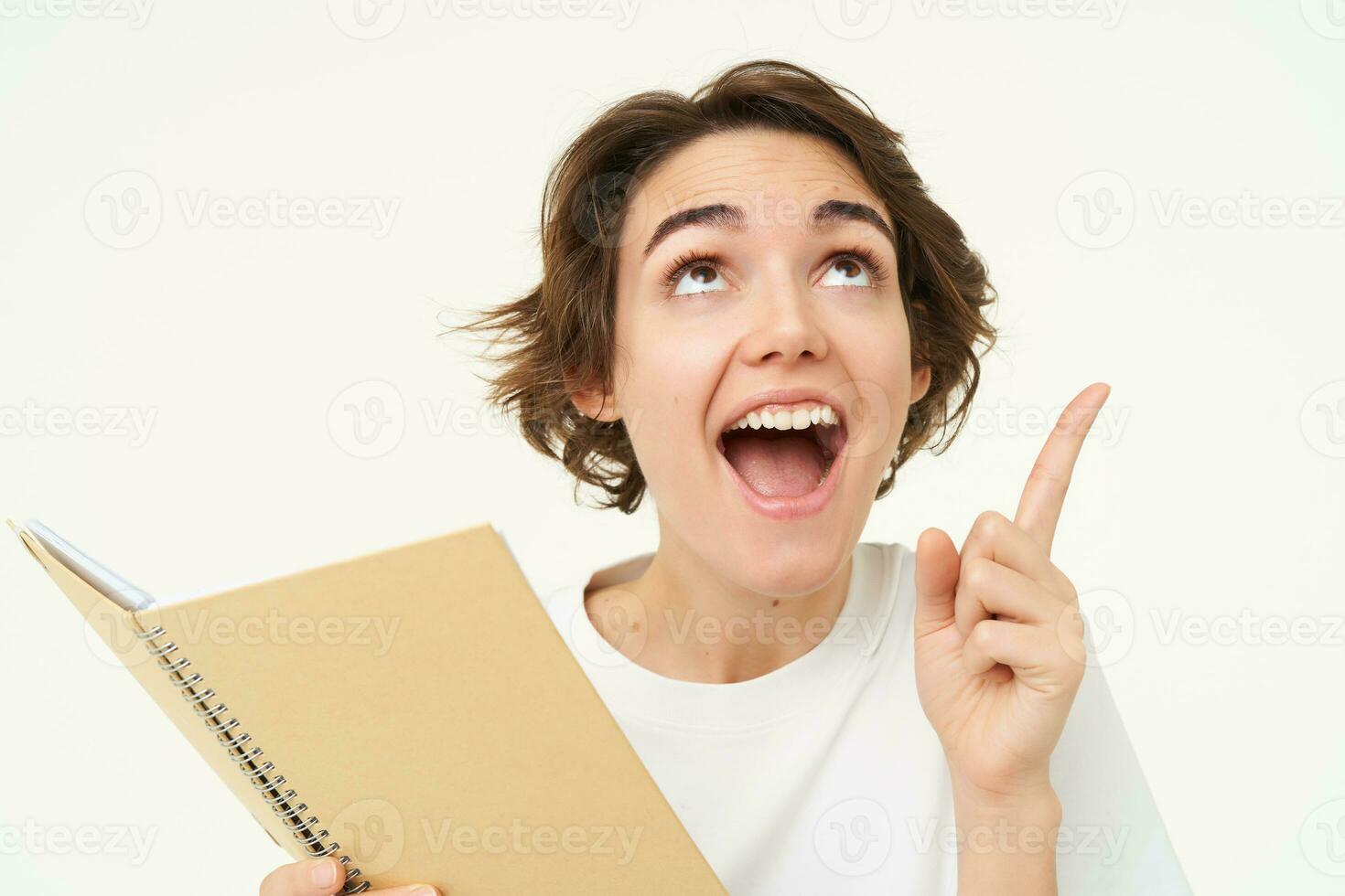 Enthusiastic brunette woman with diary, holding planner book, pointing at upper right corner and smiling with excitement, standing over white background photo