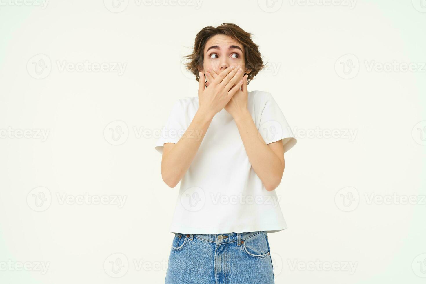 Portrait of woman with shocked, surprised face, covers her mouth with hands and gasps in amazement, stands over white background photo